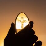 A silhouetted hand holds a crucifix against a vibrant sunset sky, symbolizing faith and spirituality.