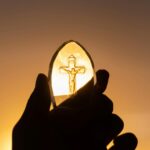 A silhouetted hand holds a crucifix against a vibrant sunset sky, symbolizing faith and spirituality.